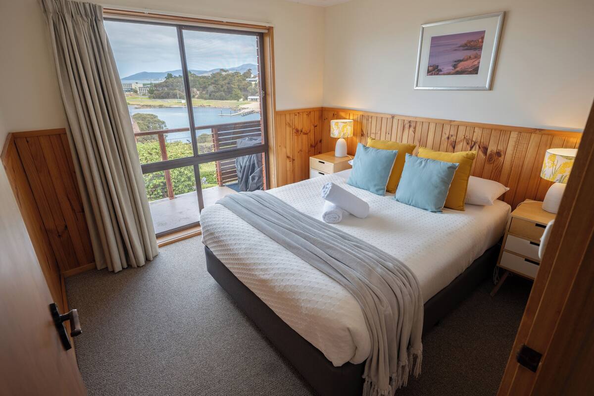 A bedroom with a large bed, wooden headboard, two bedside tables with lamps, a view of a lake and mountains through a sliding door, and a framed picture on the wall.