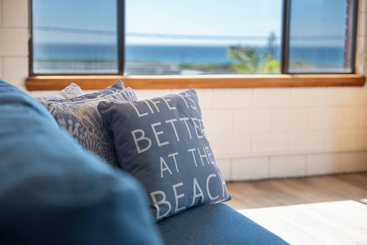 A view of a room with a large window overlooking the sea, a blue sofa with patterned cushions, and a pillow with the text "LIFE IS BETTER AT THE BEACH".
