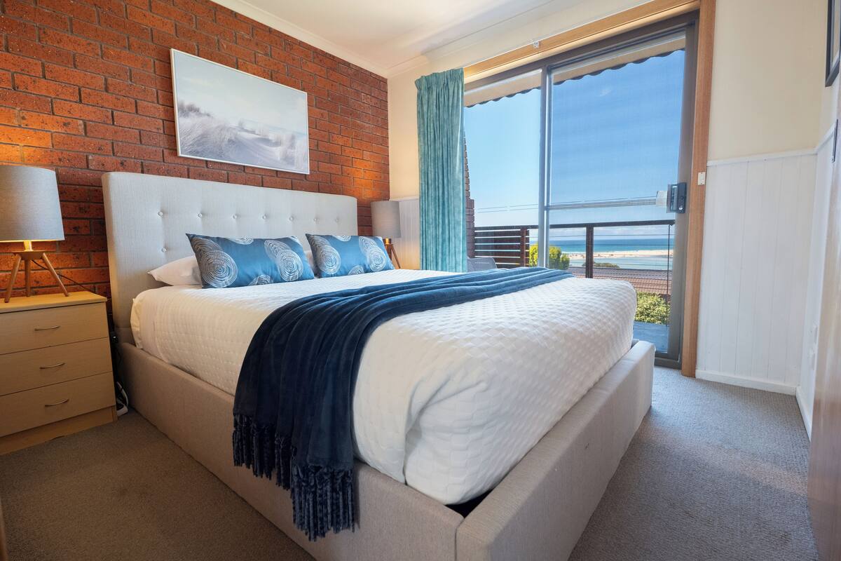 A bedroom with a large bed, a nightstand, a lamp, a view of the beach through a sliding door, and a brick wall.