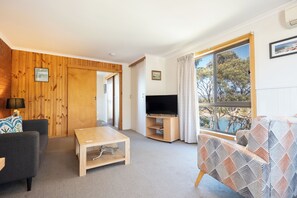 A living room with a wooden wall, a TV, a sofa, a coffee table, and a large window with a view of trees. - Blue Seas Holiday Villas (Scamander)