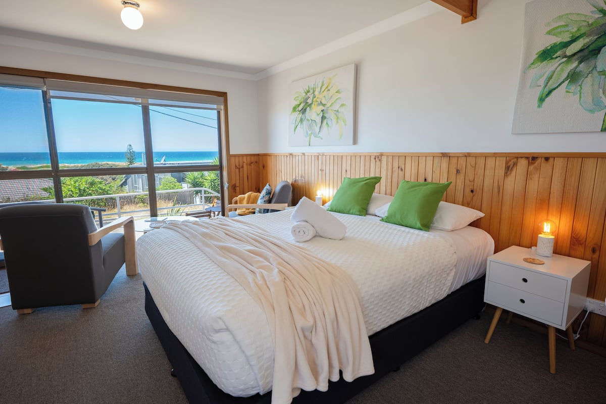 A bedroom with a large bed, a nightstand, a chair, and a view of the ocean through a large window.