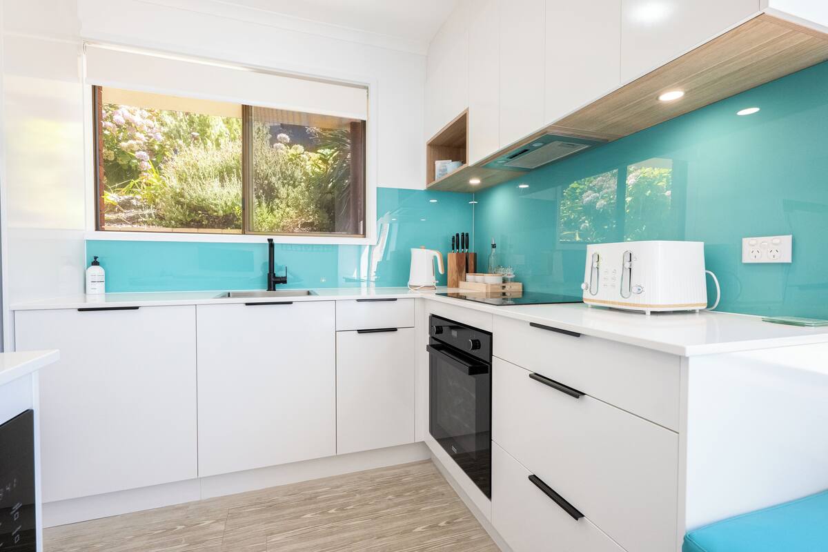 A modern kitchen with white cabinets, a black oven, a window with a garden view, and a teal backsplash.
