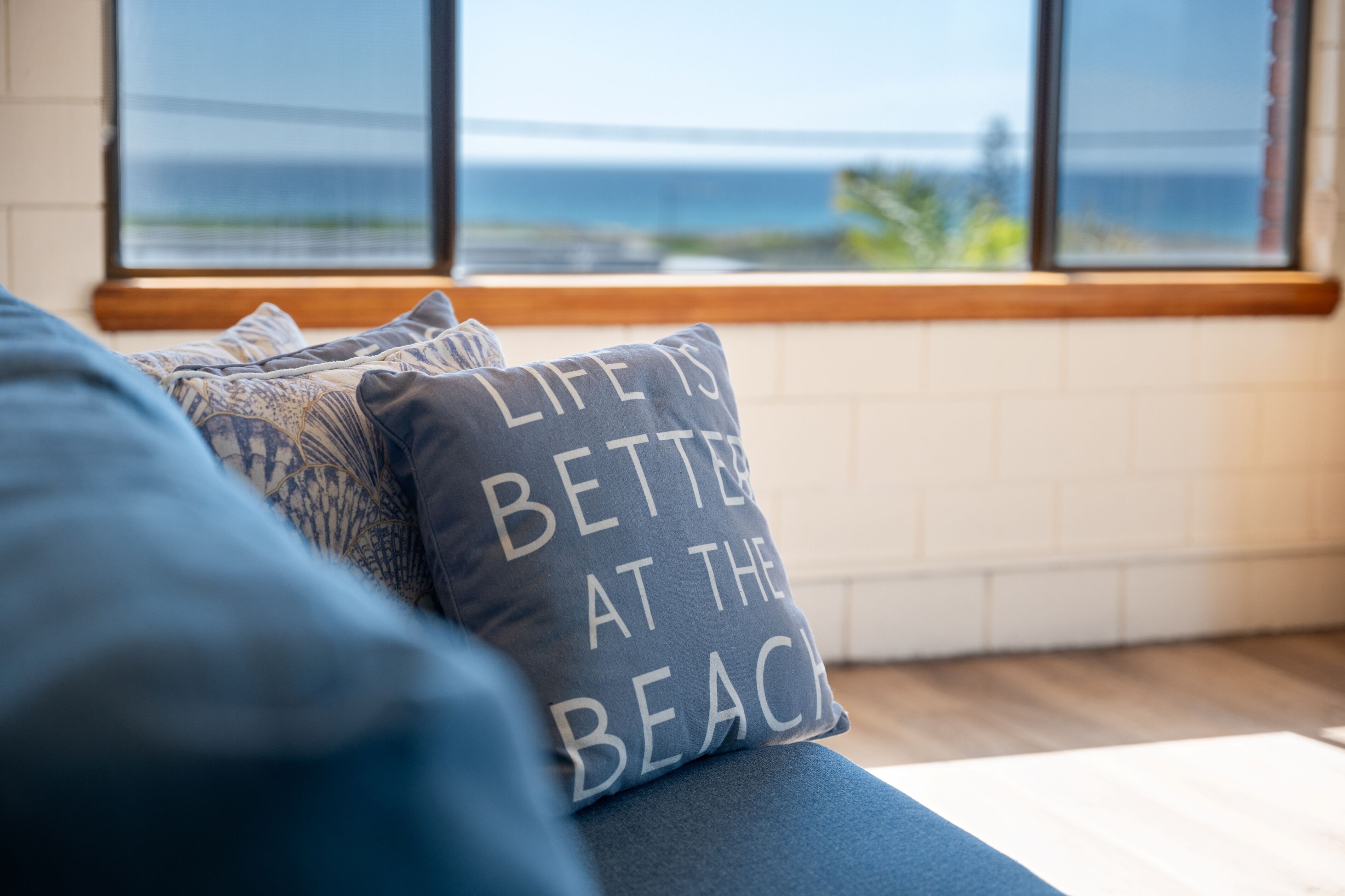 Ein Zimmer mit Blick auf das Meer durch große Fenster, ein blaues Sofa mit gemusterten Kissen und ein Kissen mit dem Aufdruck 'Life is better at the beach'.