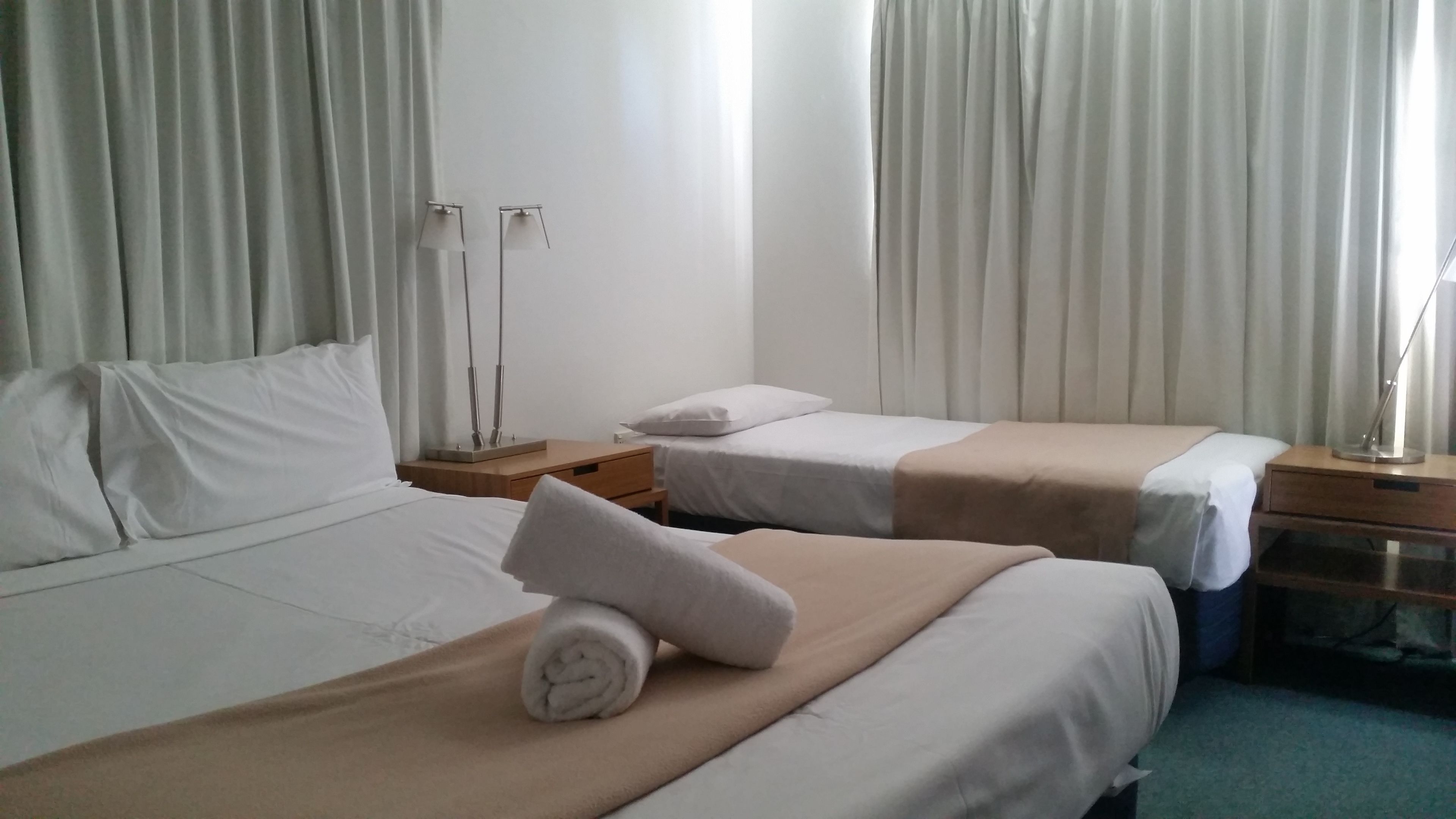 A hotel room with two beds, a nightstand, a lamp, and a window with curtains.
