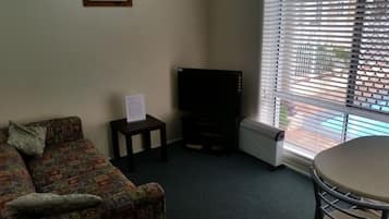 Una sala de estar con un sofá estampado, un televisor de pantalla plana sobre un mueble, una mesita con un letrero y una ventana con persianas.