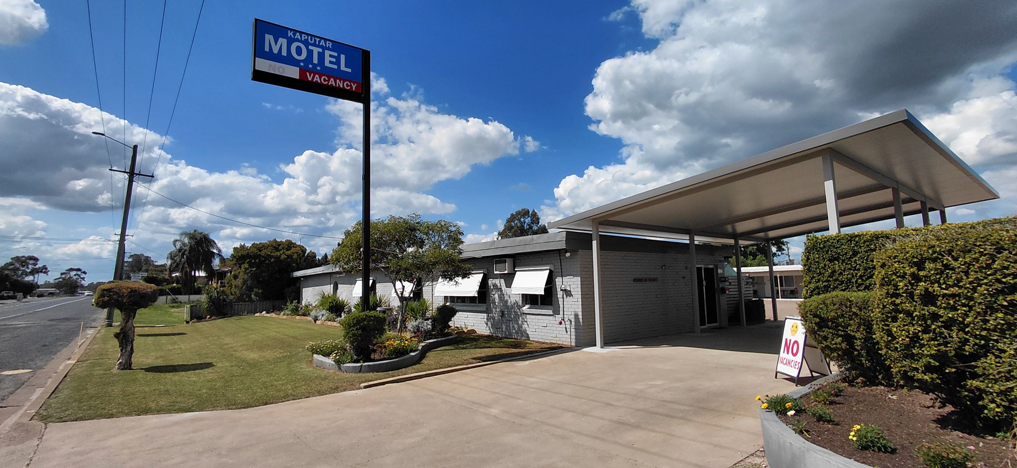 Un motel con área de estacionamiento techada, un letrero que indica habitaciones agotadas y cielo despejado.