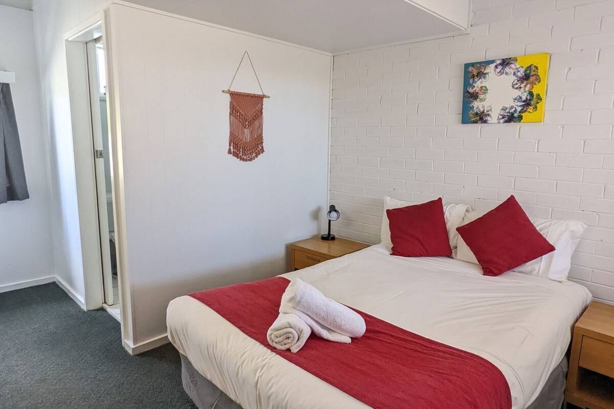 A bedroom with a bed, a nightstand, a macrame wall hanging, and a floral artwork.
