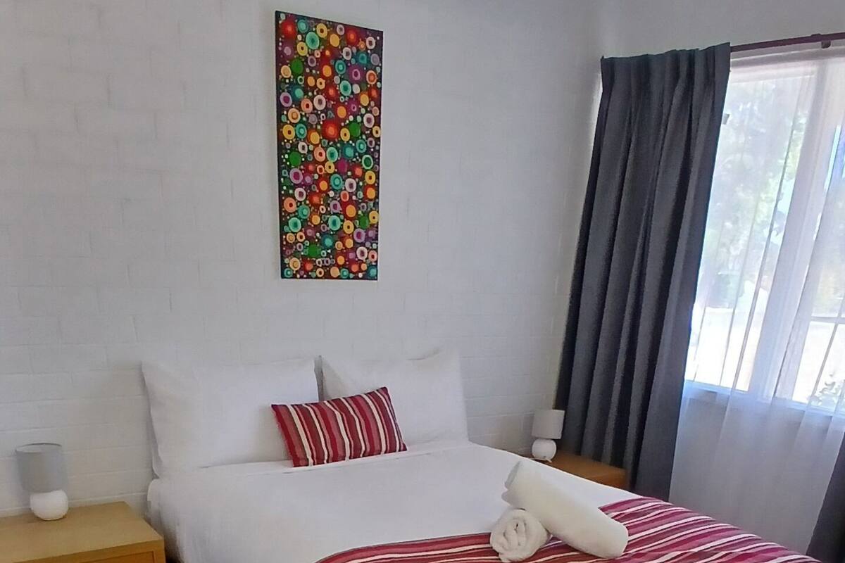A neatly arranged hotel room with a large bed, a bedside table with a lamp, a colorful abstract painting on the wall, and a window with curtains.