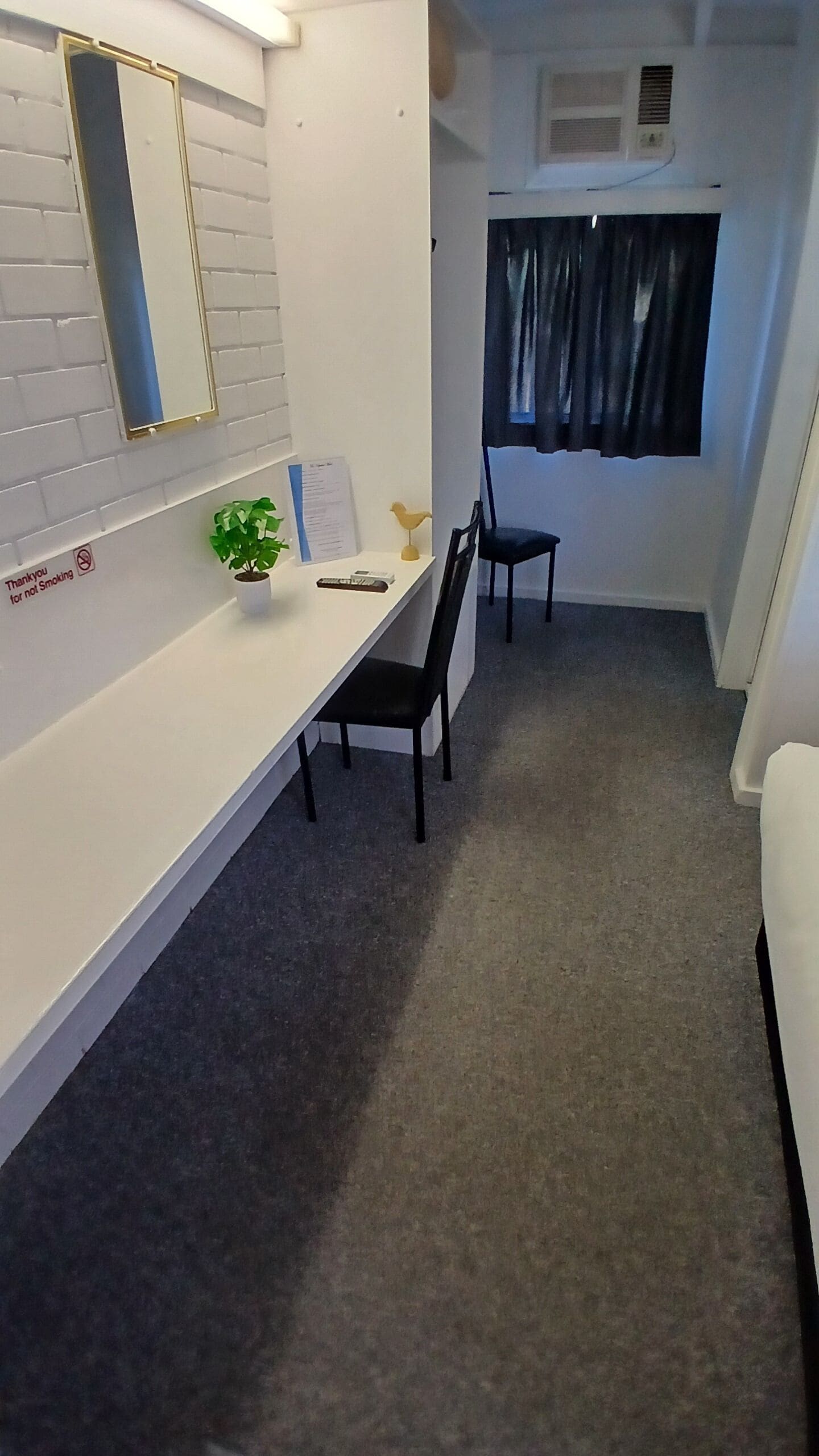 Ein kleiner, sauberer Raum mit einem weißen Schreibtisch, zwei schwarzen Stühlen, einer kleinen Pflanze und einem Fenster mit Vorhängen.
