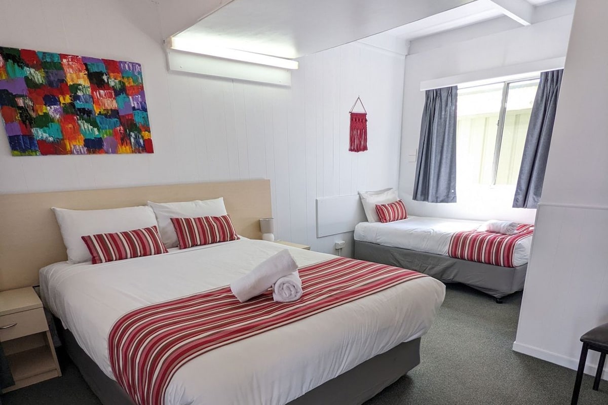 A hotel room with two beds, a large abstract painting on the wall, a window with curtains, and a small table.