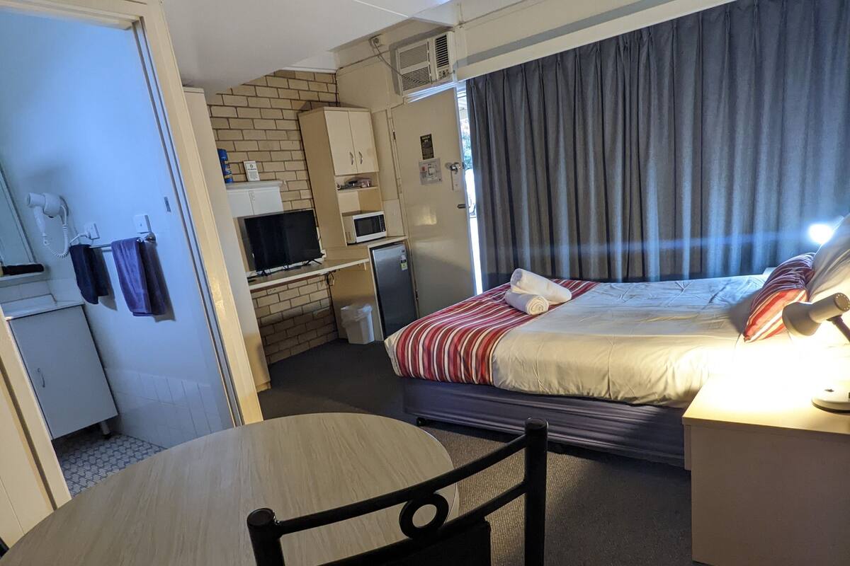 A compact hotel room with a bed, a small kitchenette, a dining table with chairs, and a separate bathroom area.