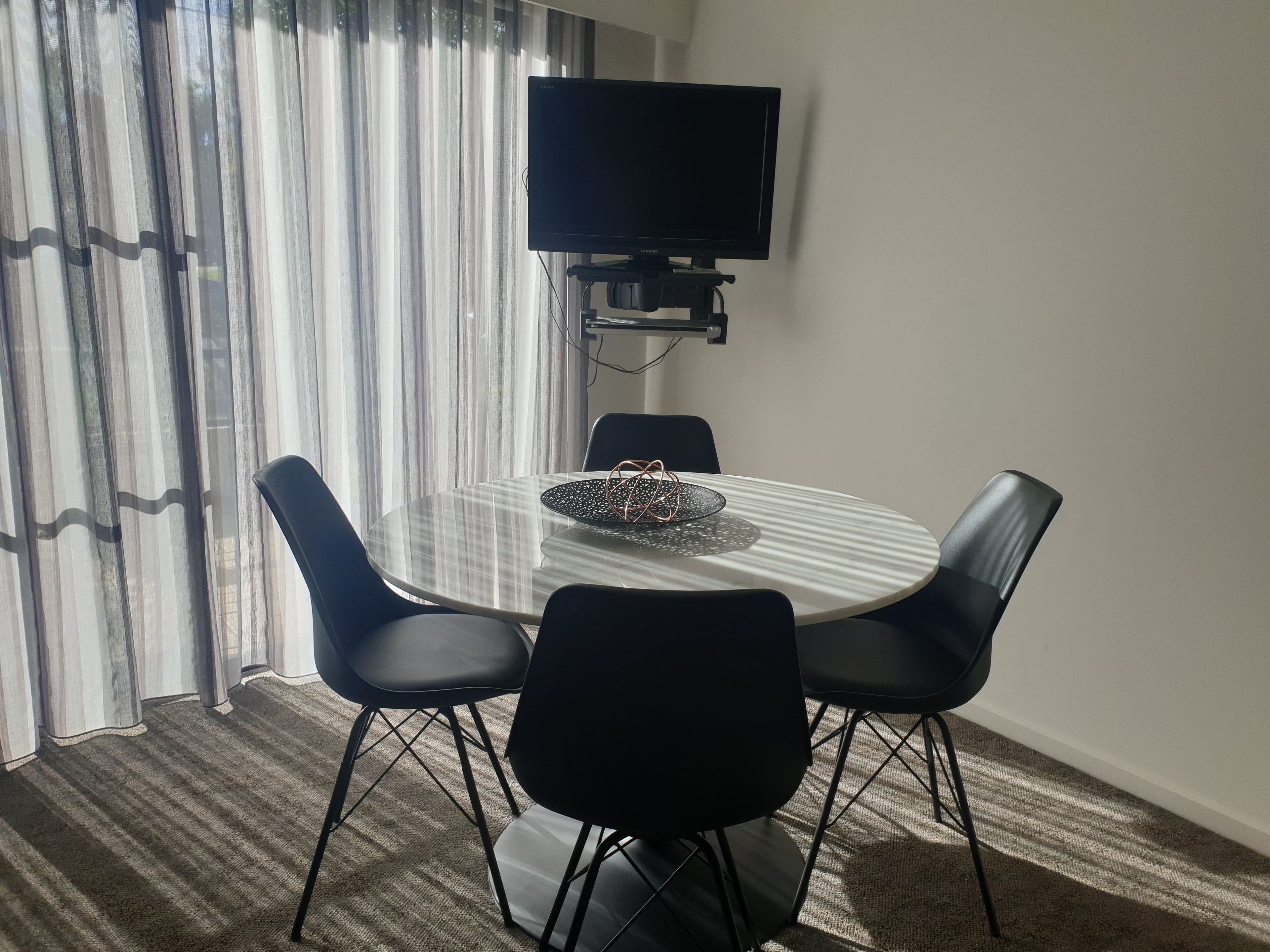 A modern dining area with a round table, four black chairs, a television mounted on the wall, and sheer curtains.