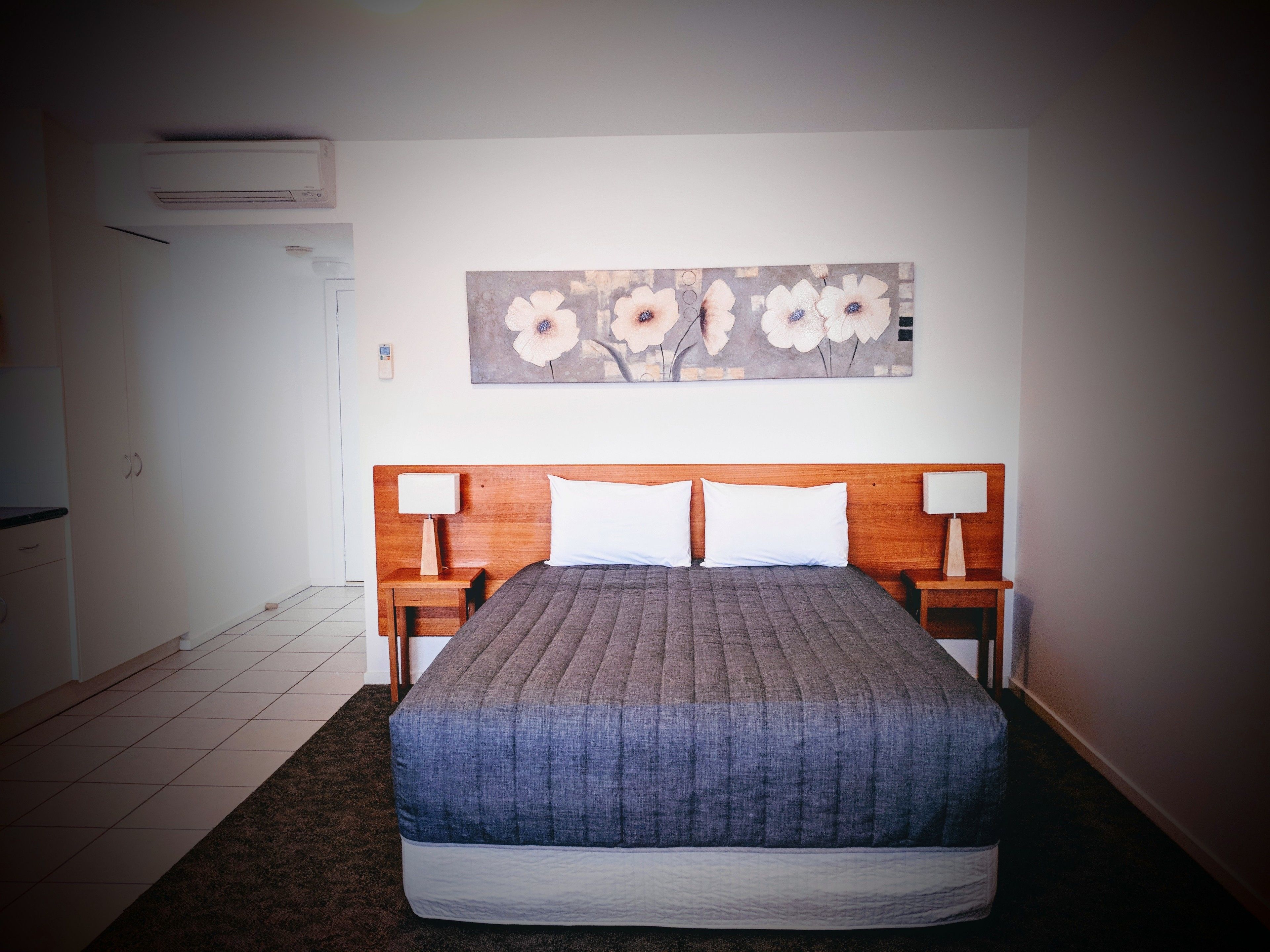 Una habitación de hotel con una cama grande, dos mesitas de noche con lámparas, un aire acondicionado instalado en la pared y un cuadro floral sobre la cama.