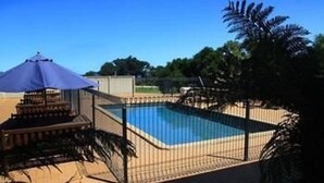 Une piscine entourée d’une clôture, un espace de repos à l’ombre et un ciel d’un bleu limpide.