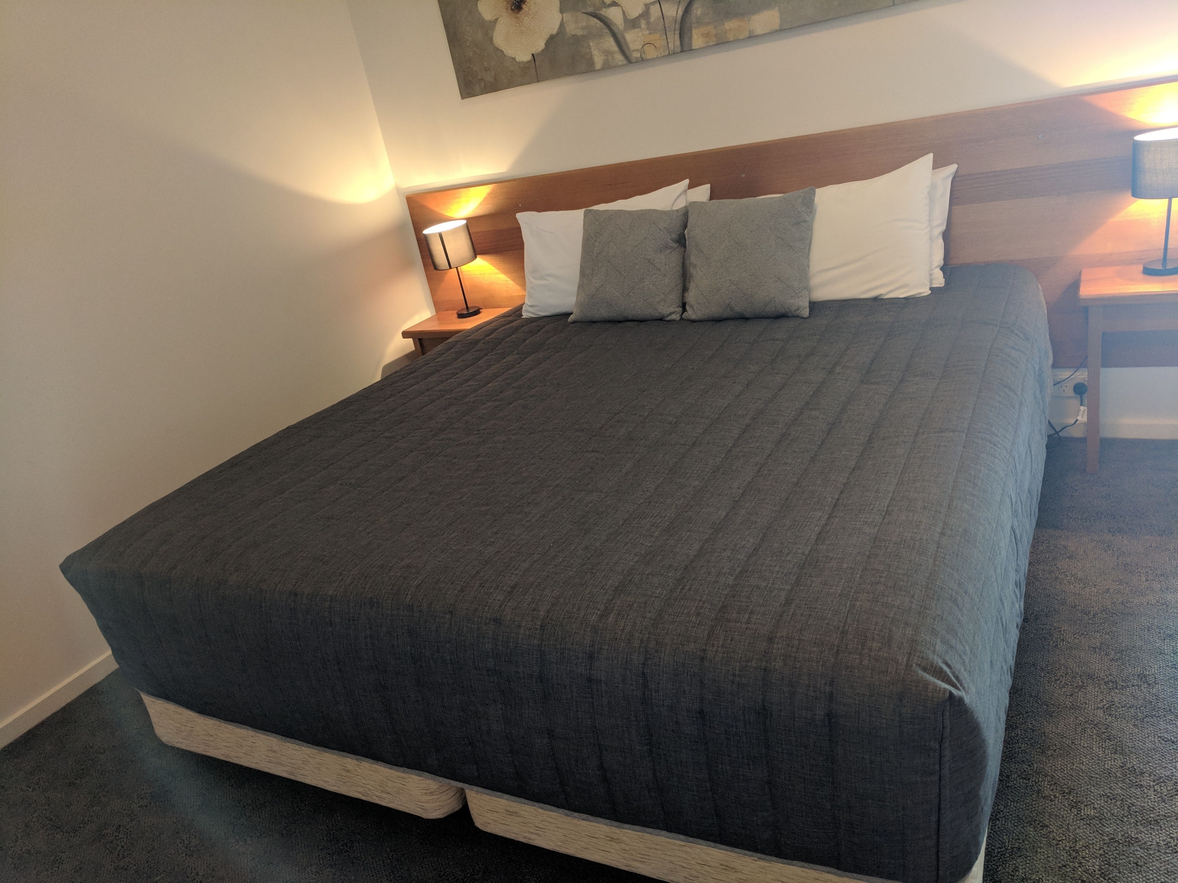 Cama bem arrumada com colcha cinza escura e travesseiros, cabeceira de madeira e duas luminárias de cabeceira.