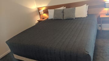 Cama bem arrumada com colcha cinza escura e travesseiros, cabeceira de madeira e duas luminárias de cabeceira.