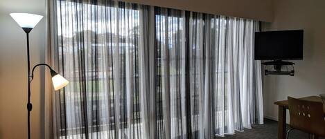A hotel room with a large window, a flat-screen TV mounted on the wall, a sofa, a coffee table, and a lamp.