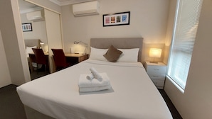 Habitación de hotel con una cama bien hecha, un escritorio con silla, una lámpara de noche y una ventana con persianas.
