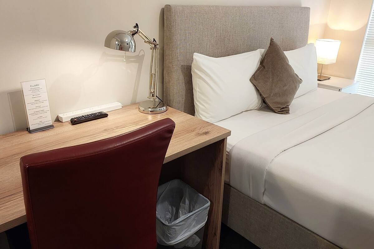 Une chambre d’hôtel avec un bureau en bois, une chaise rouge, un lit avec des oreillers blancs et bruns, une lampe et un panneau indiquant les équipements de la chambre.