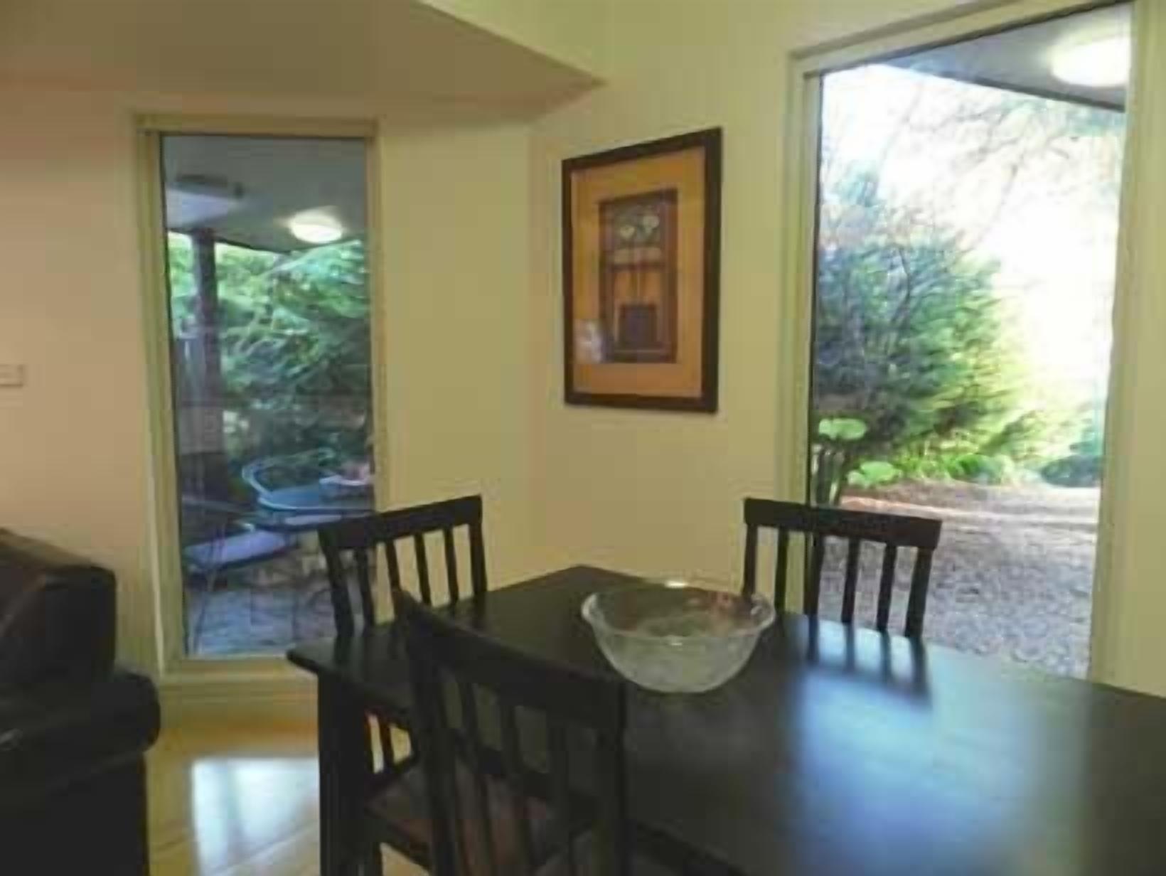 A dining area with a table and chairs, a framed picture on the wall, and a view of greenery outside.