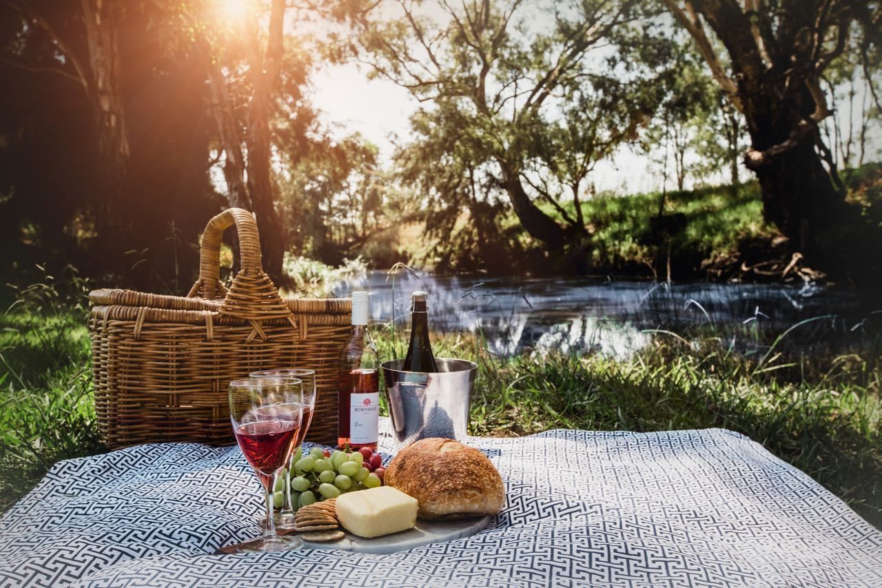 A picnic setup with a wicker basket, wine bottle, glasses, grapes, cheese, and bread on a patterned blanket by a body of water.