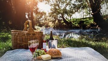 A picnic setup with a wicker basket, wine bottle, glasses, grapes, cheese, and bread on a patterned blanket by a body of water.