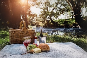 A picnic setup with a wicker basket, wine bottle, glasses, grapes, cheese, and bread on a patterned blanket by a body of water. - Evanslea Luxury Boutique Accommodation (Mudgee)