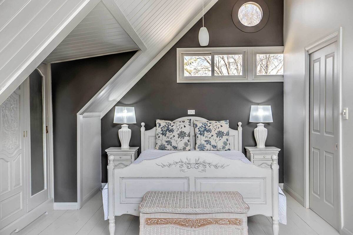 Une chambre à coucher avec un lit blanc, deux tables de chevet avec des lampes, une fenêtre donnant sur un beau paysage et une porte.