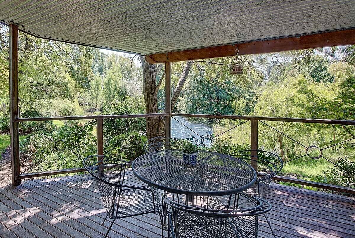 A covered outdoor deck with a round metal table and chairs, surrounded by a wooden railing, overlooking a lush green landscape.