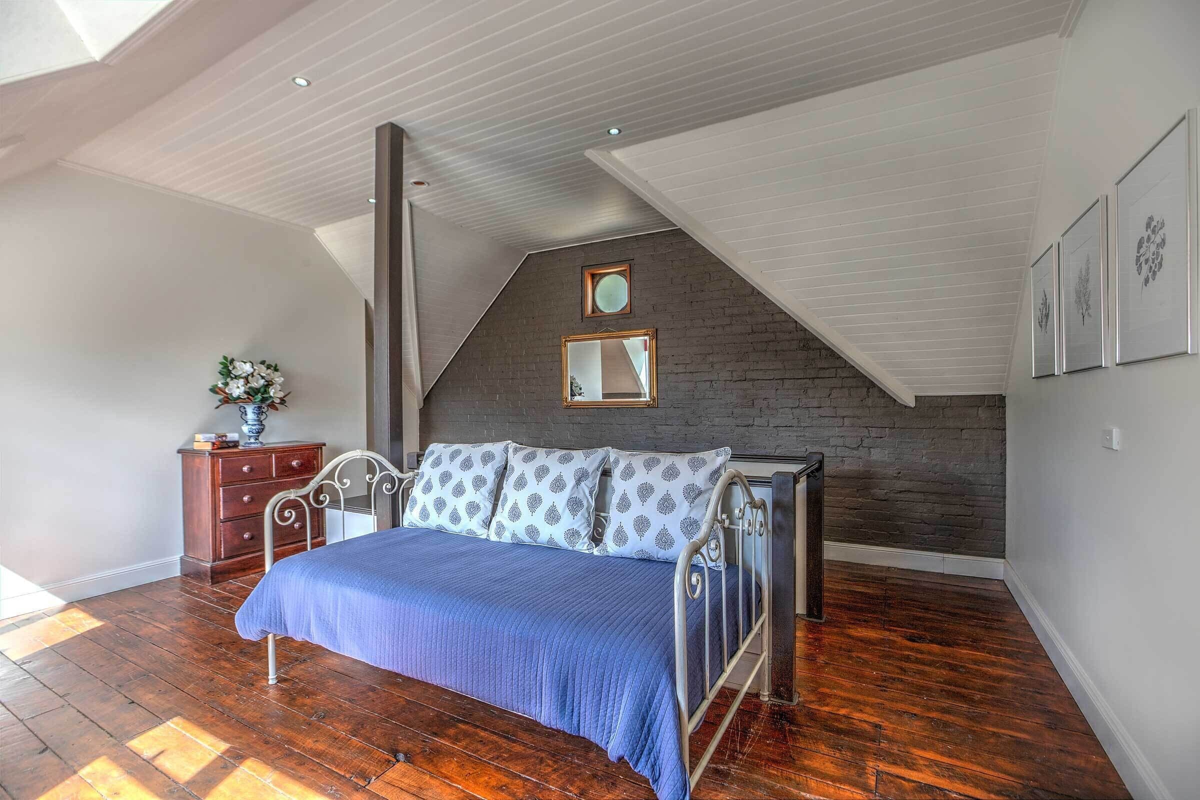 A bedroom with a sloped ceiling, a bed with a blue bedspread, a dresser, and a brick accent wall.