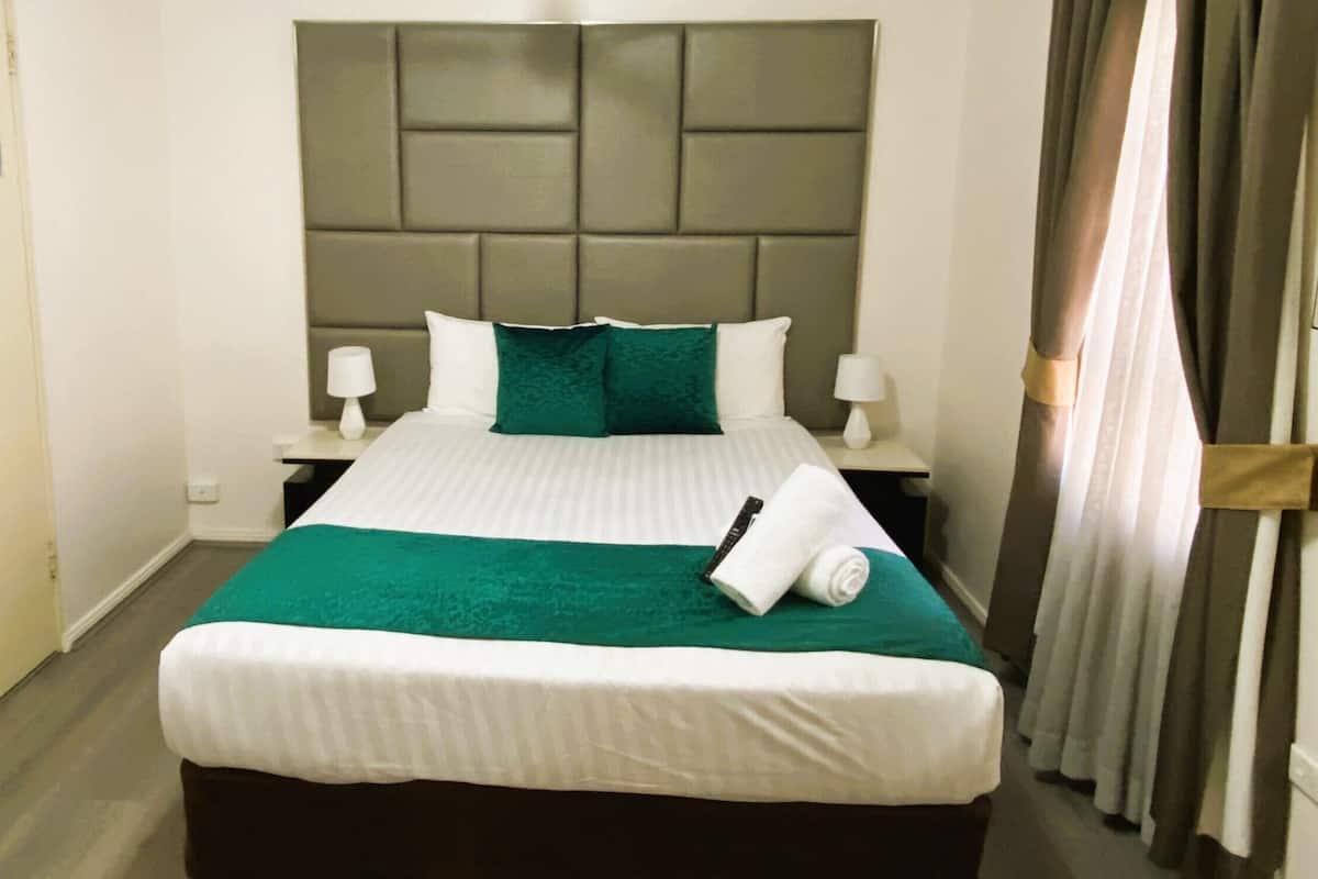 A neatly made bed with a green bedspread and white pillows, a headboard with a geometric pattern, two bedside lamps, and a window with curtains.