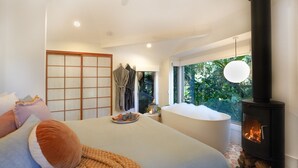 A bedroom with a large bed, a built-in bathtub, a wood-burning stove, and a view of greenery through the window. - Lovestone Cottages (Montville)