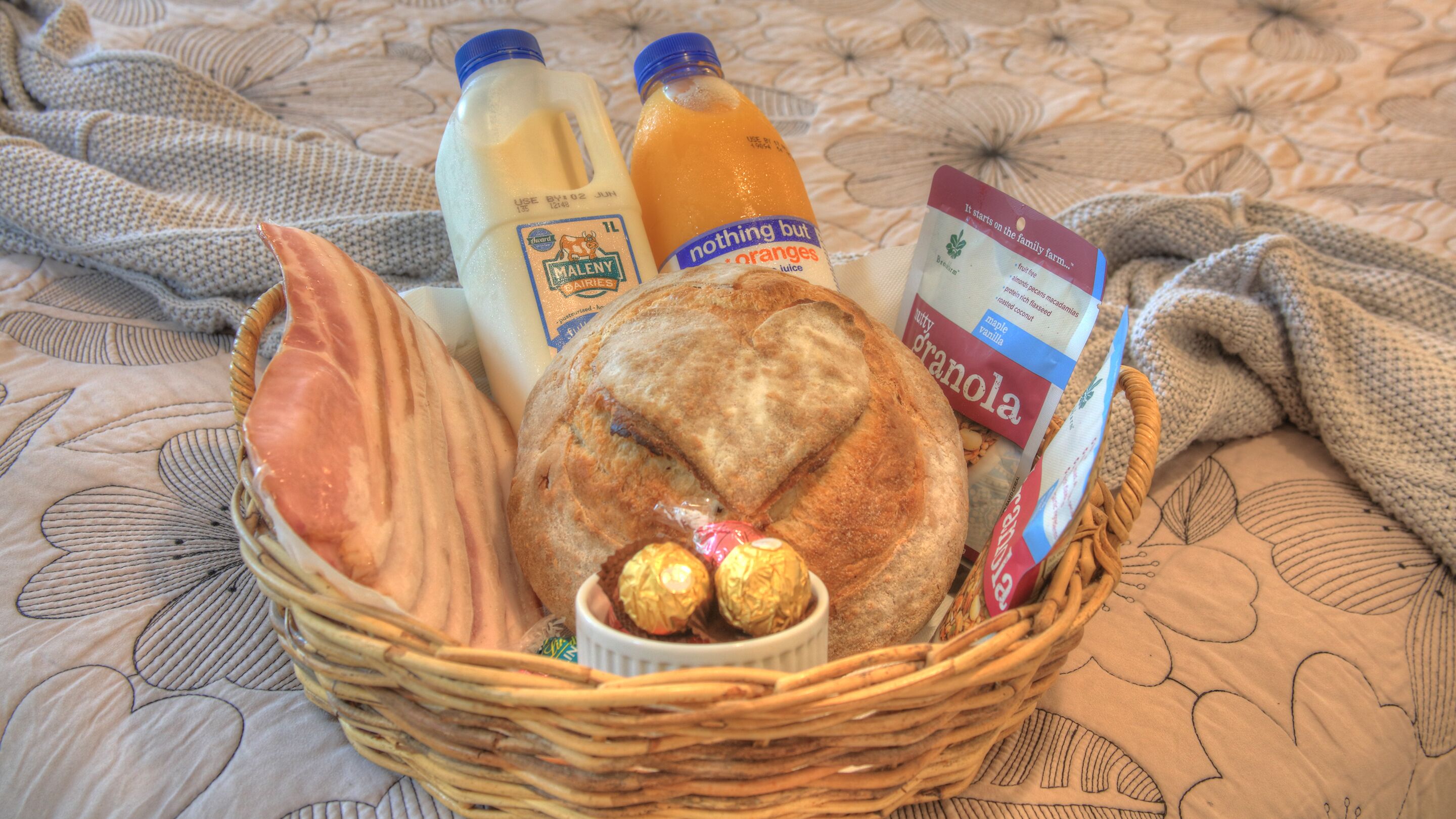 A basket containing bread, milk, orange juice, and granola on a patterned bedspread.