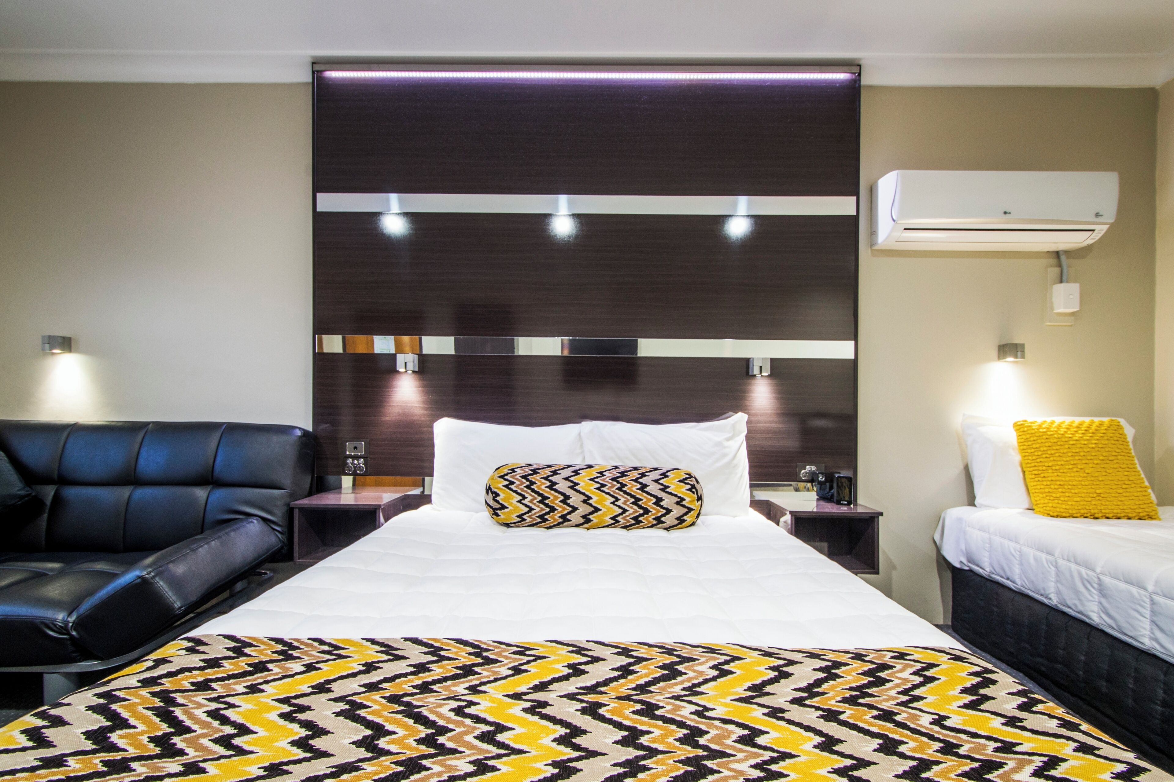 Habitación de hotel con cama, sofá de cuero negro, mesita de noche, aire acondicionado y lámpara de pared.