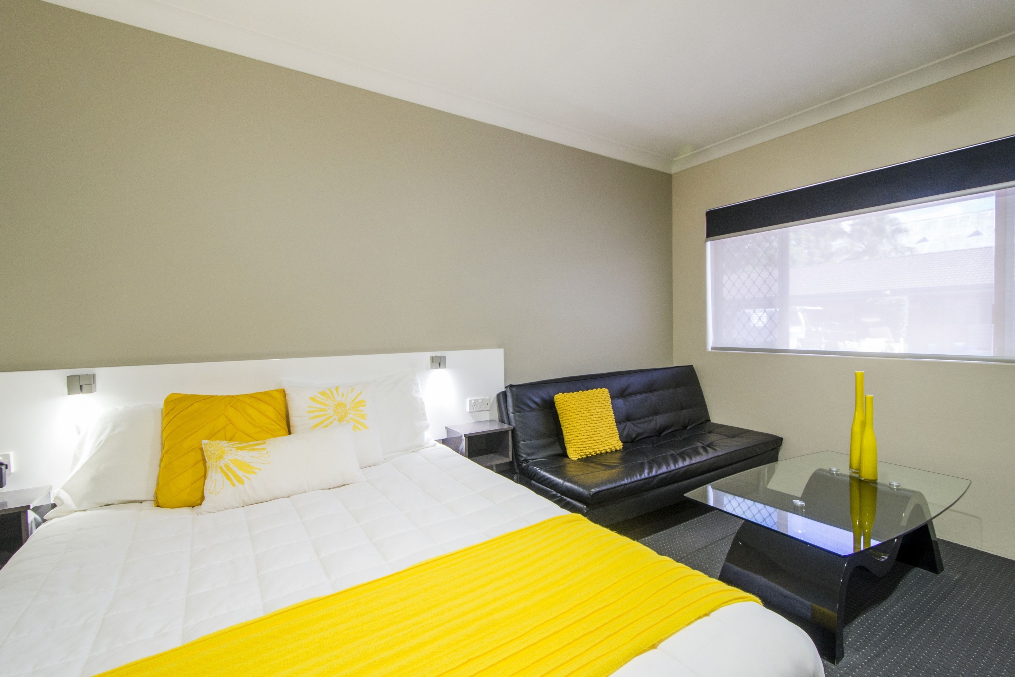 A hotel room with a bed, a black leather sofa, a glass coffee table, and a window with blinds.