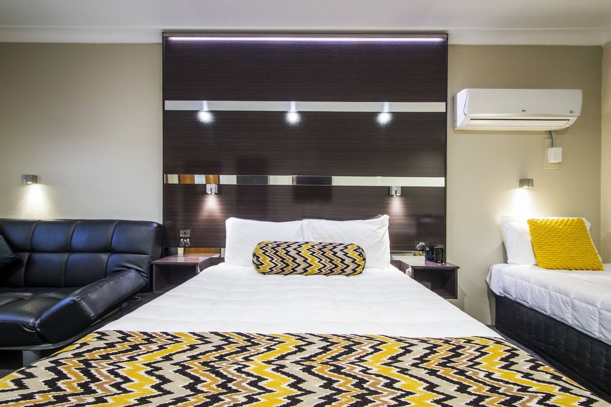 A hotel room with a large bed, a black leather sofa, a bedside table, an air conditioner, and a wall-mounted light fixture.