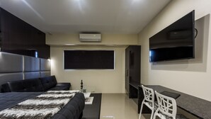 A modern hotel room with a large bed, a desk with two chairs, a flat-screen TV mounted on the wall, and an air conditioning unit.
