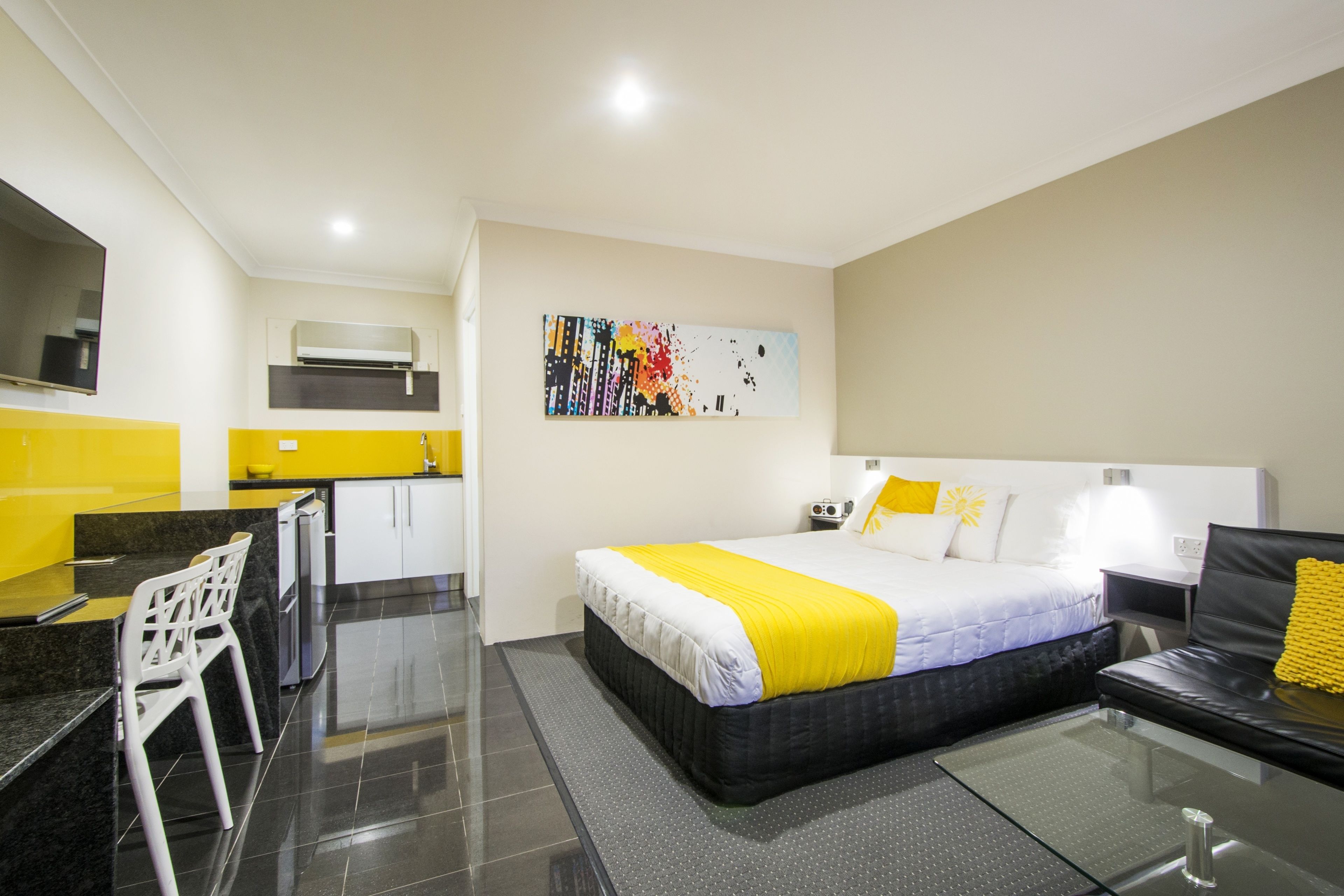 A modern hotel room with a bed, a desk, a chair, and a kitchenette.