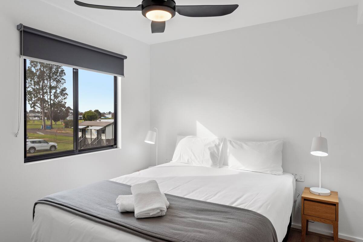 Habitación de hotel moderna con una cama grande, un ventilador de techo, una mesita de noche con lámpara y una ventana con vistas al exterior.
