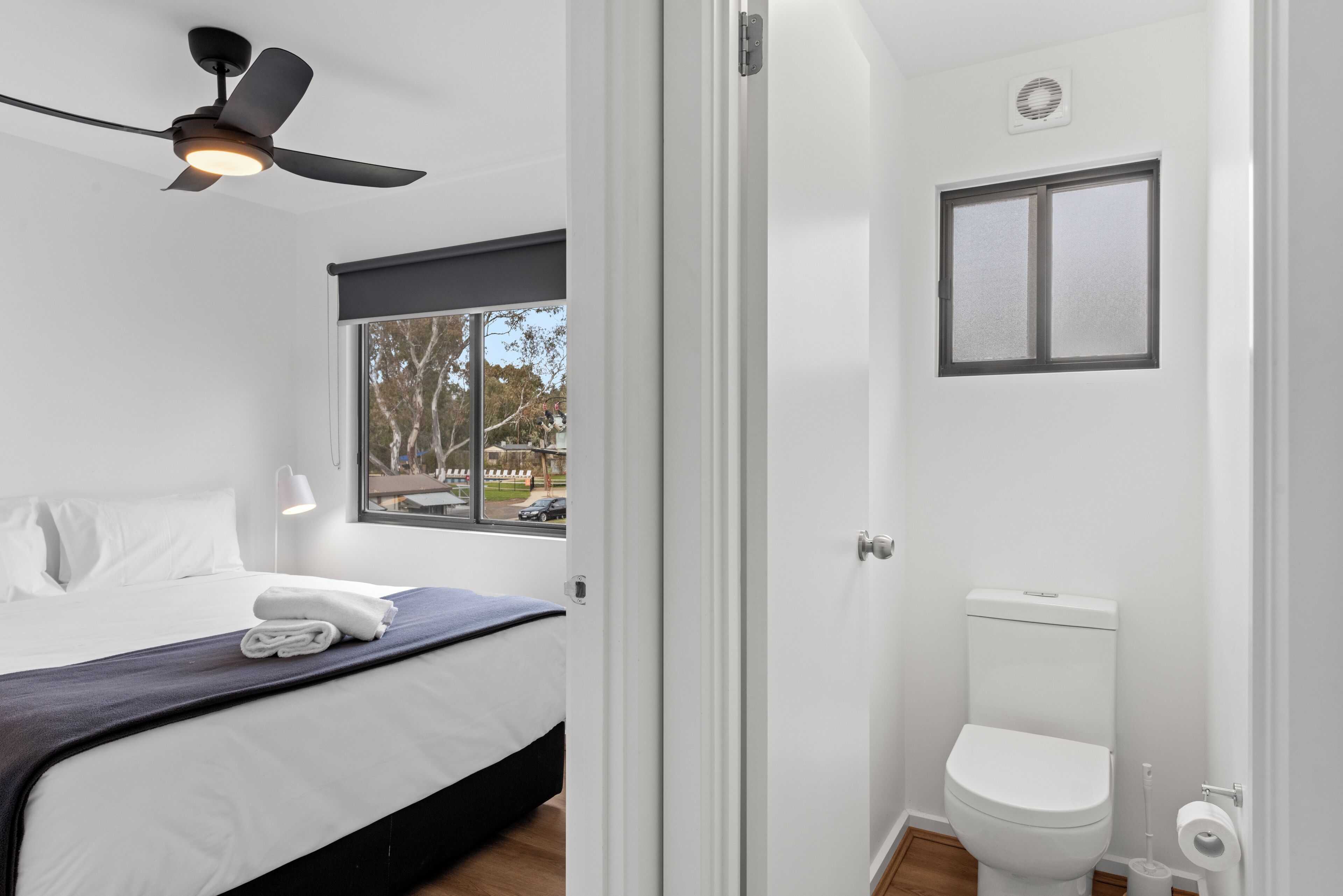 Ein Schlafzimmer mit Bett, Deckenventilator, Fenster mit Aussicht und einem Badezimmer mit Toilette und Fenster.