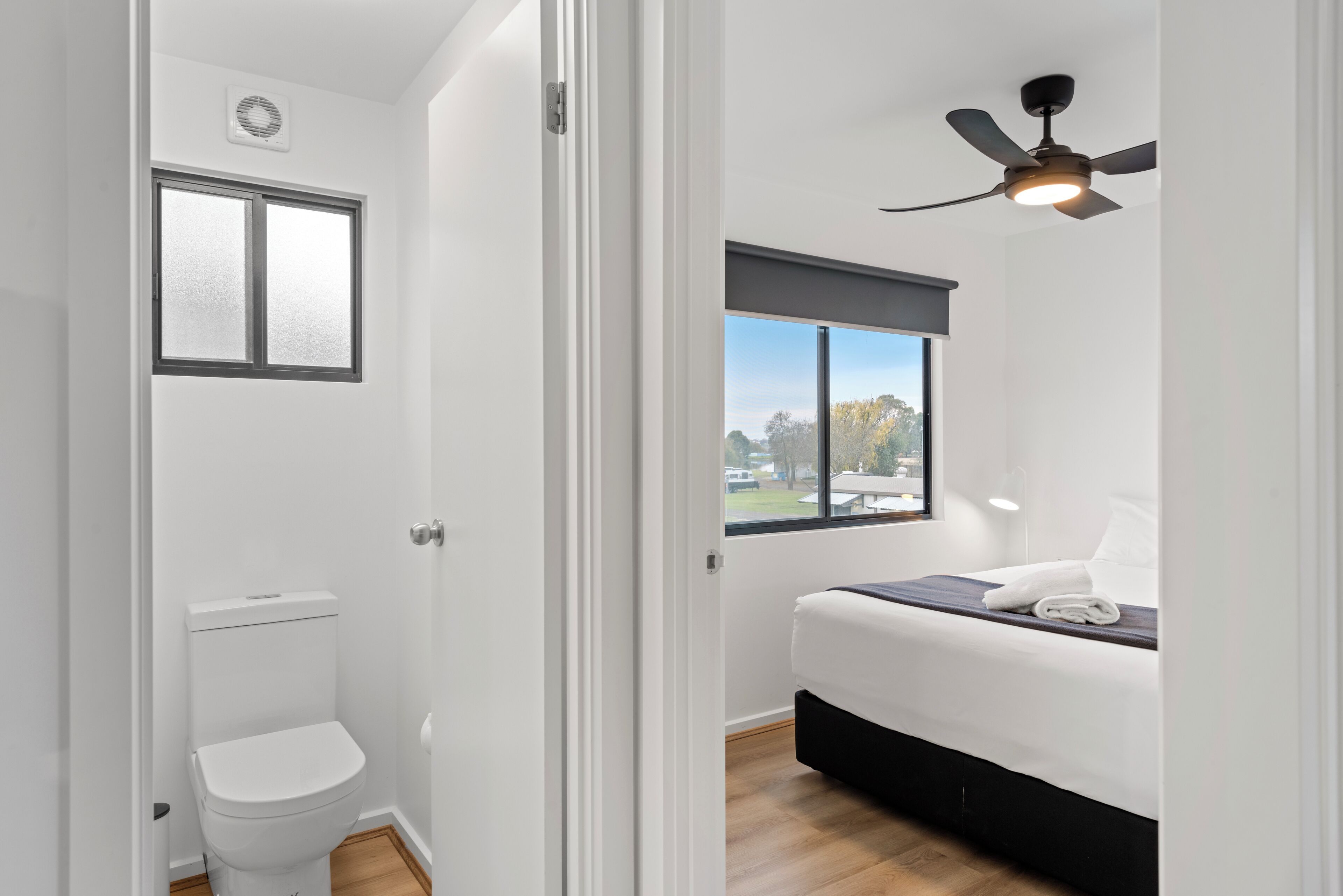 A modern bedroom with a bed, a window with a view, a ceiling fan, and a white toilet.