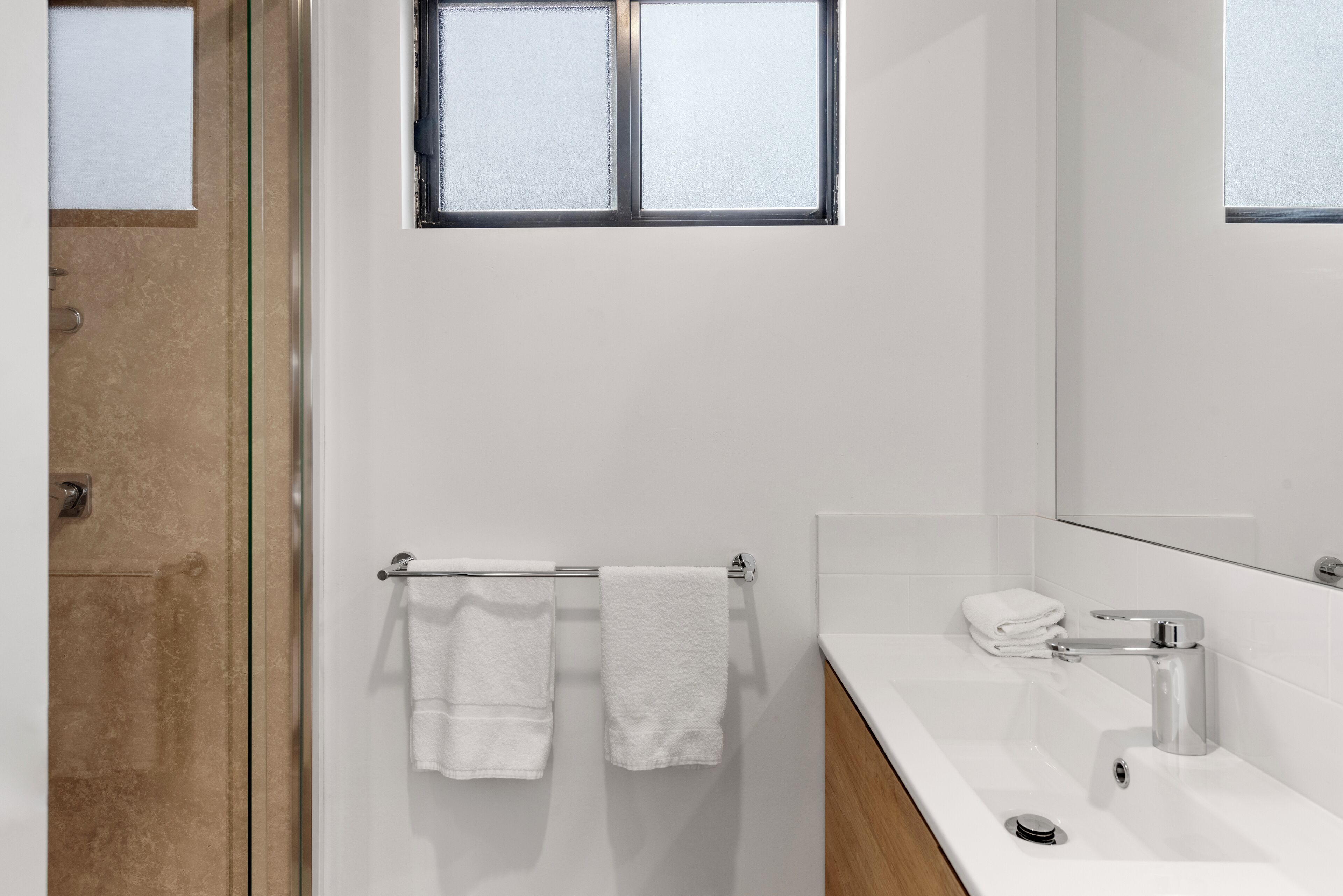 Een moderne badkamer met een glazen douchedeur, een witte wastafel met een chromen kraan en een handdoekrek met twee witte handdoeken.