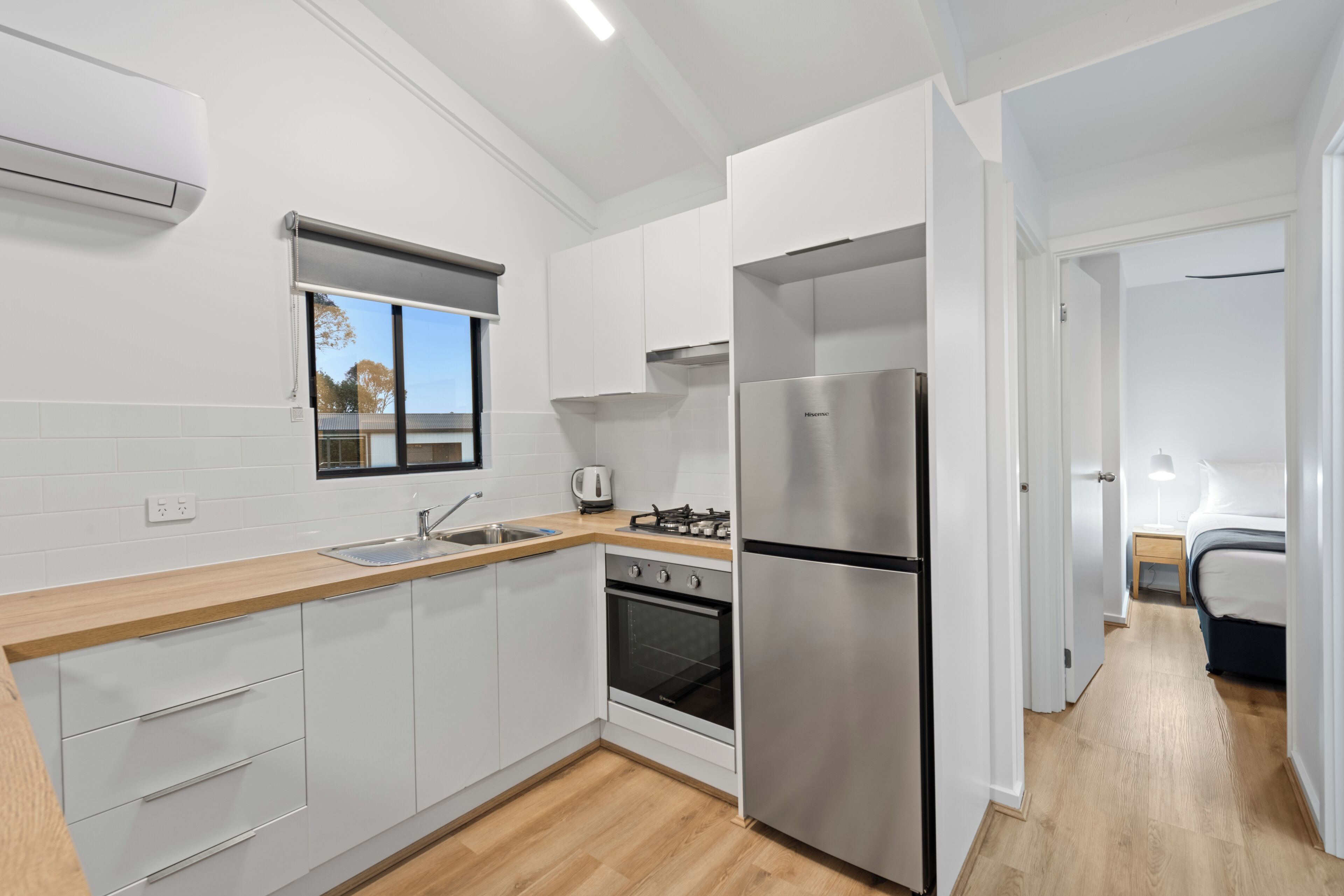 A modern kitchen with white cabinets, a stainless steel refrigerator, a built-in oven, and a window with blinds.