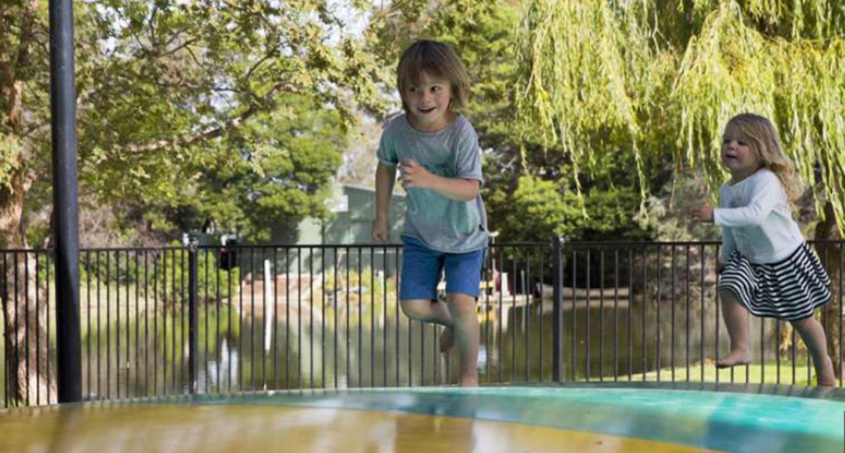 Dos niños jugando en un trampolín en un parque con una valla y árboles al fondo.