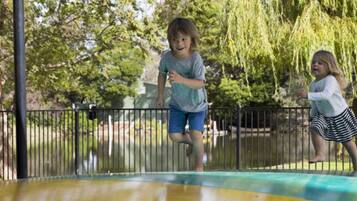 Dos niños jugando en un trampolín en un parque con una valla y árboles al fondo.