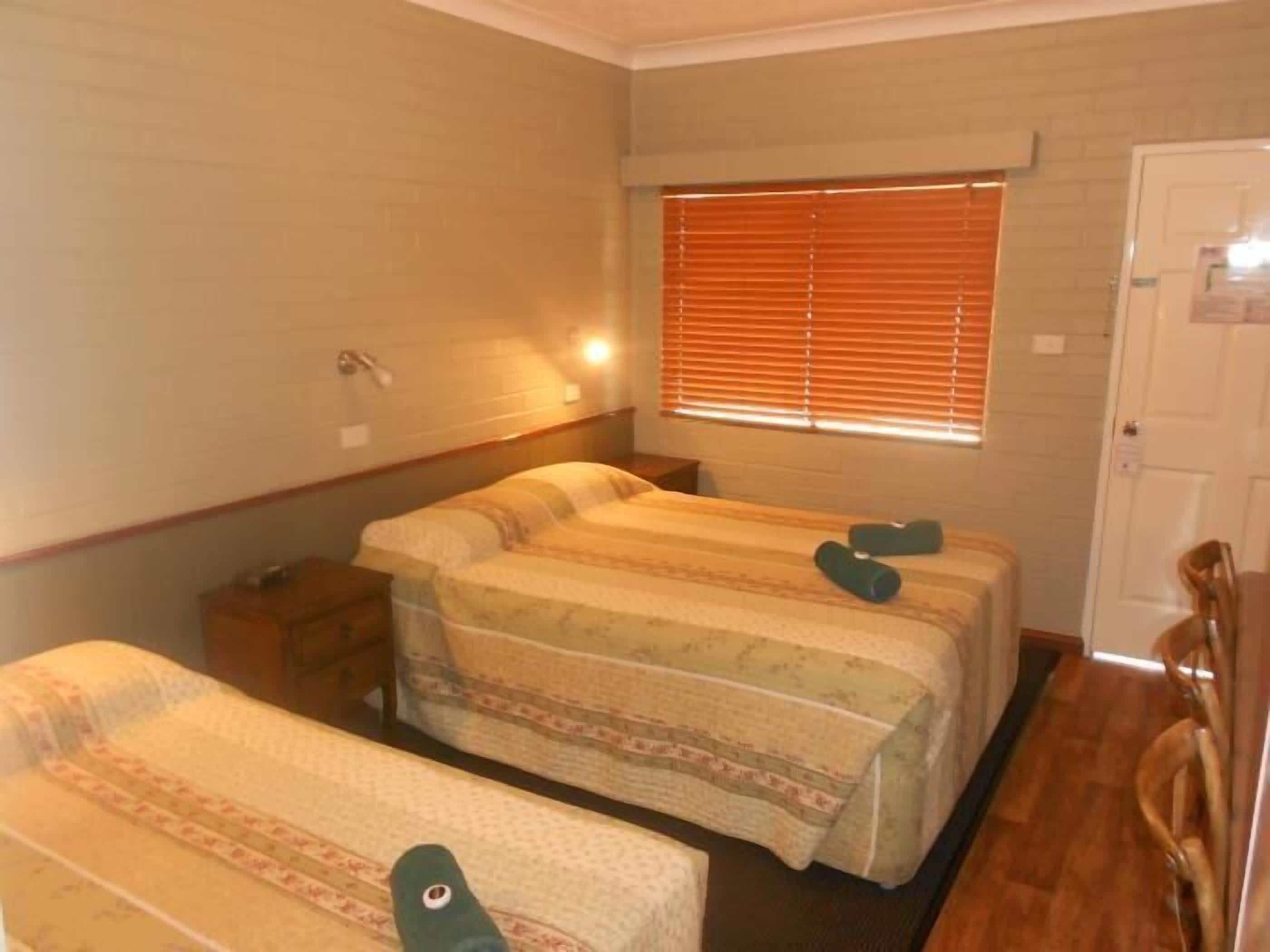 A room with two beds, a wooden floor, a window with blinds, a door, and a small table.