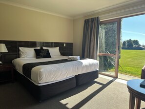 A hotel room with a large bed, a nightstand, a lamp, and a view of a grassy area through a glass door.