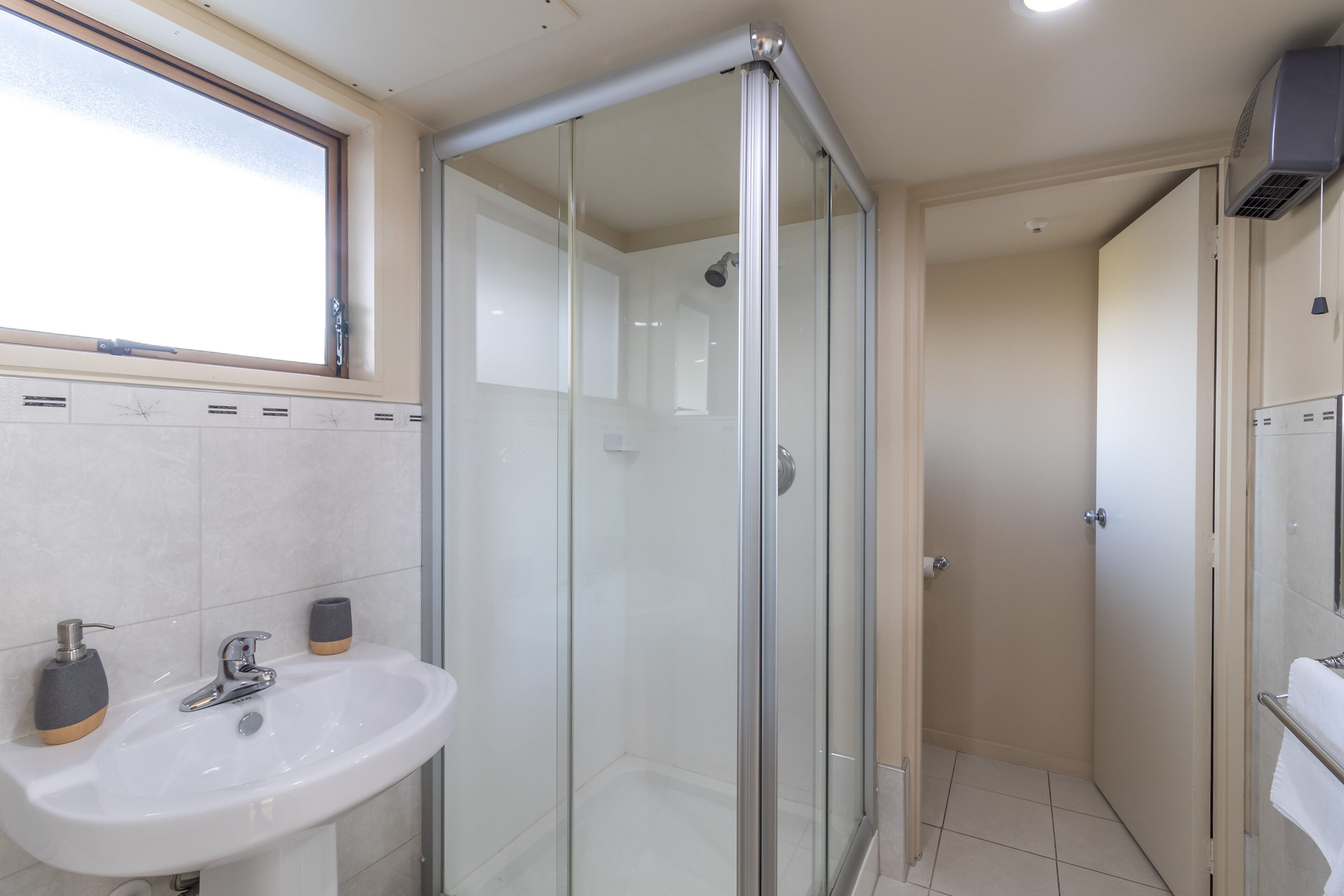 Un baño moderno con cabina de ducha de vidrio, un lavamanos con dos dispensadores de jabón y una ventana sobre el lavamanos.