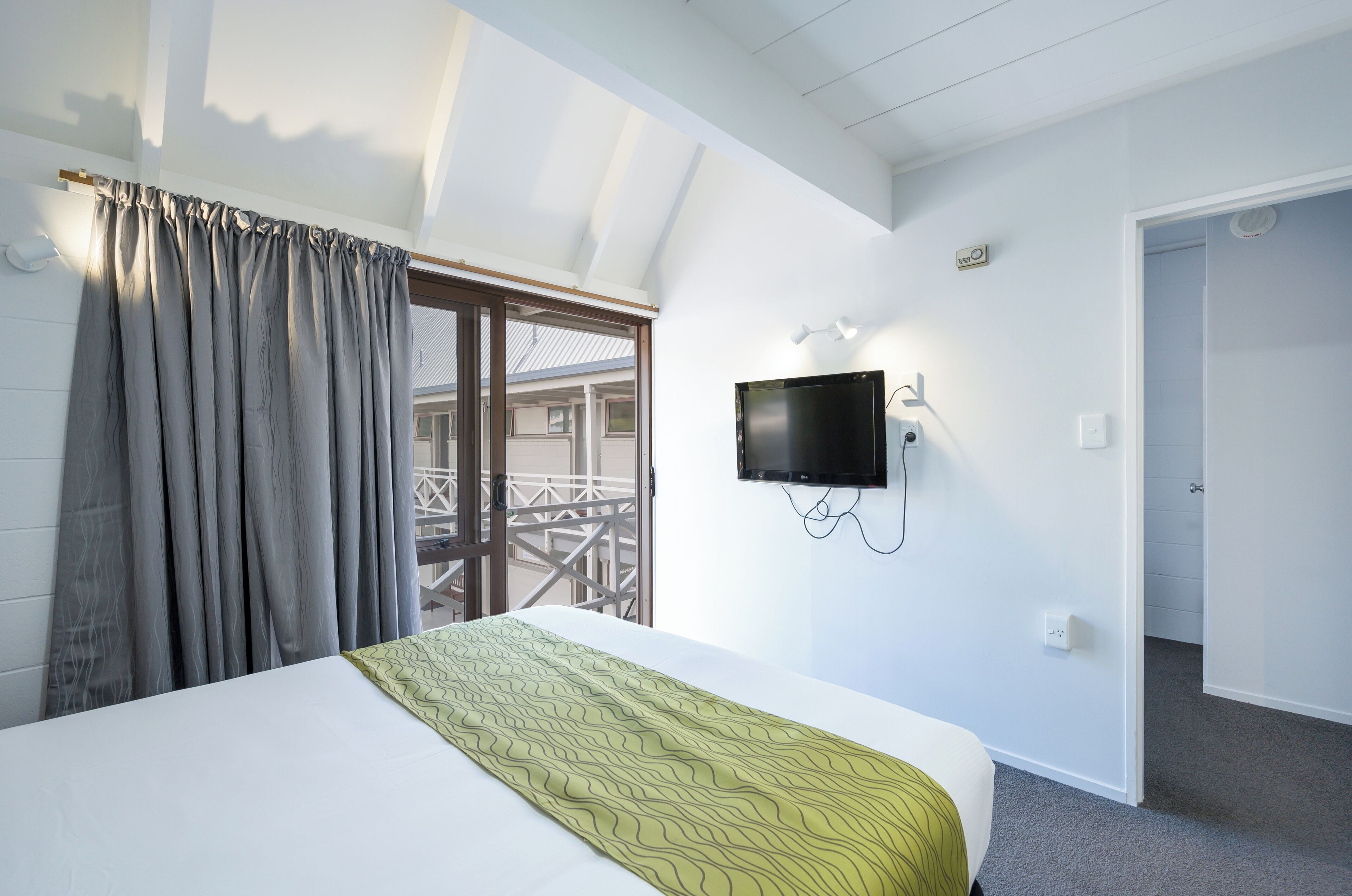 Een hotelkamer met een bed, een aan de muur bevestigde televisie, een balkon met een hekwerk en een deur naar een andere kamer.