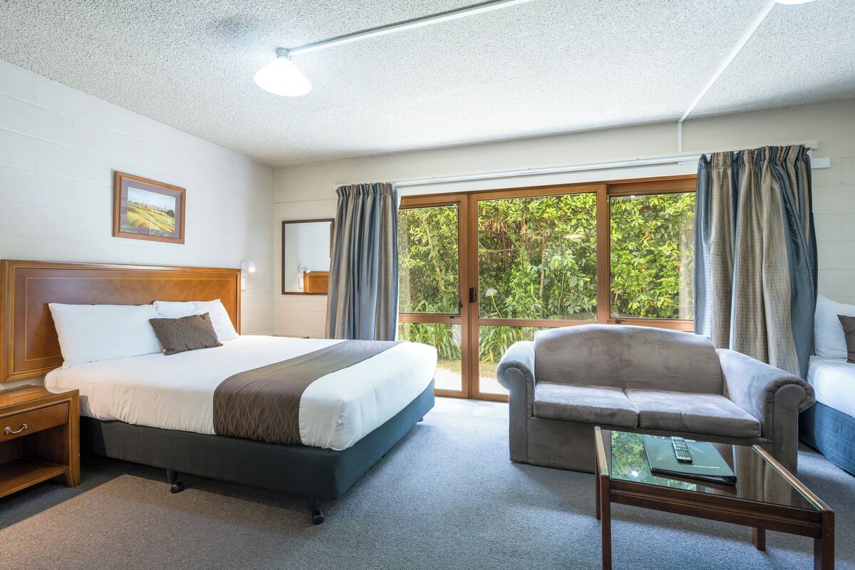 A hotel room with a large bed, a sofa, a coffee table, and a sliding glass door leading to a balcony with a view of greenery.
